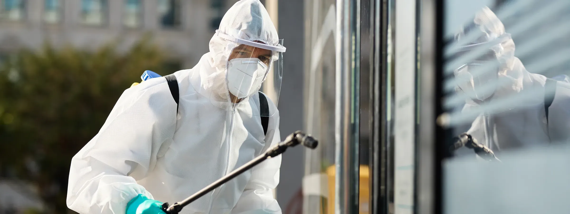 Técnico con overol blanco, careta y guantes fumigando con bomba de mano frente a la fachada de vidrio de un edificio en Bogotá